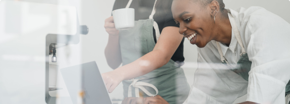 A woman leaning forward working on laptop wth another individual to her right point towards the laptop and has a mug in the opposite hand